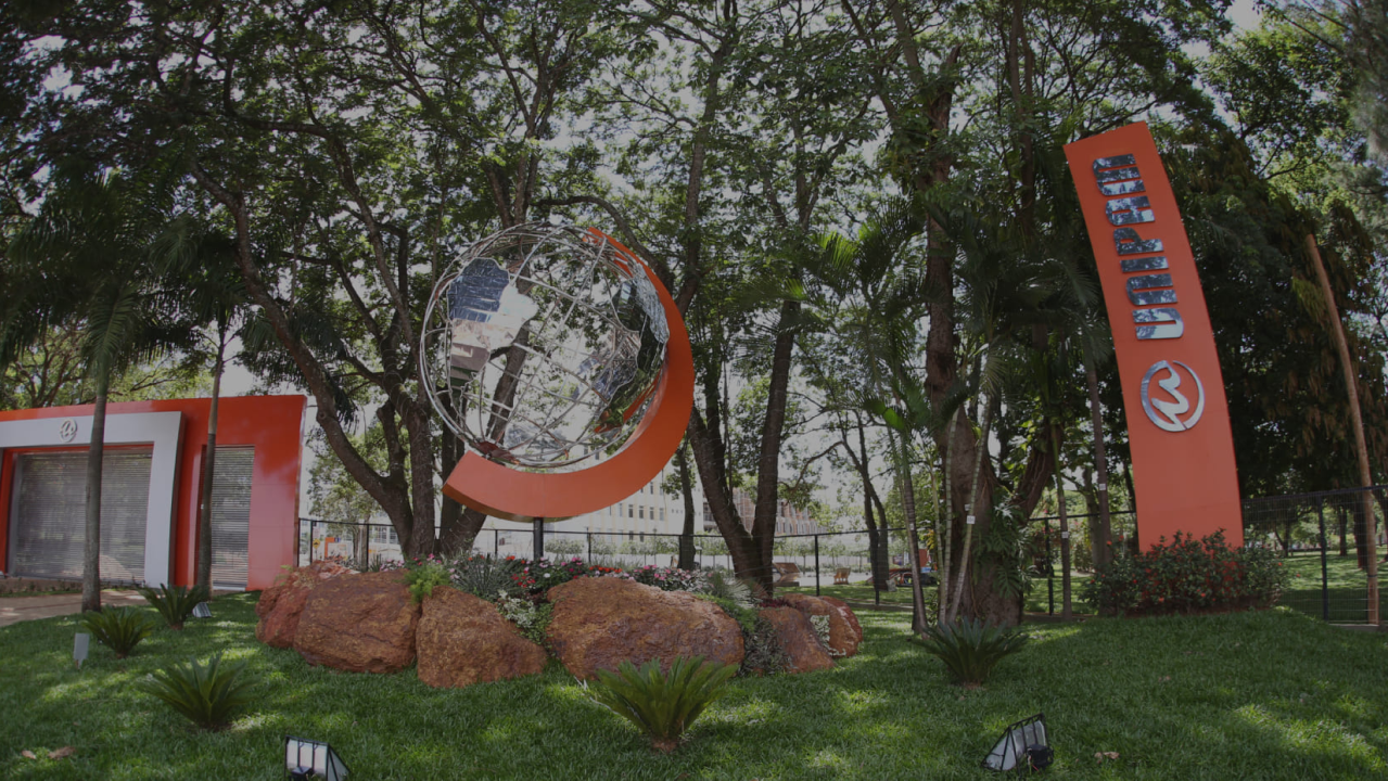 Entrada da Universidade de Patos de Minas (UNIPAM), com escultura metálica de globo terrestre rodeada por arco laranja, jardins com pedras e vegetação, e placa vertical laranja com o logotipo e nome da instituição.